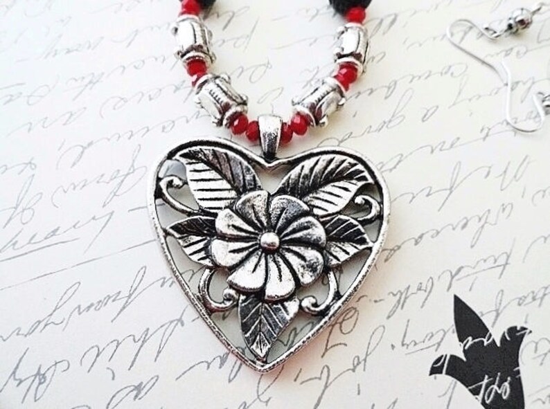 May include: A silver heart-shaped pendant necklace with a floral design. The necklace is made of red beads and silver beads.