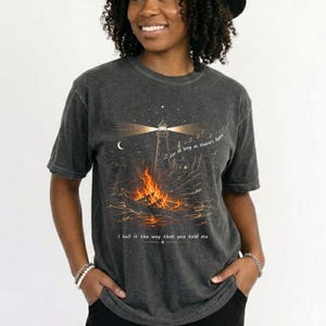 May include: A charcoal gray t-shirt featuring a graphic of a lighthouse and a burning boat on water. The text reads, "For as long as there's light" and "I tell it the way that you told me." The design includes a crescent moon and stars.