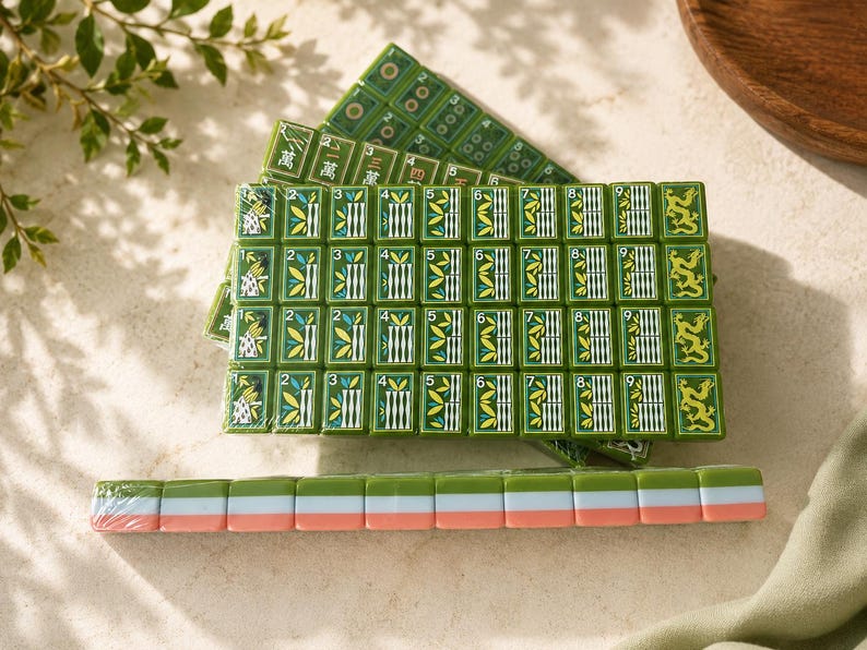 May include: A set of green mahjong tiles arranged in a rectangular grid, featuring bamboo patterns numbered one through nine, dragon symbols, and Chinese characters. A single row of tiles with green, white, and pink edges lies in the foreground.