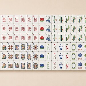 May include: A complete set of white Mahjong tiles arranged in a grid. The tiles feature red Chinese characters, colorful floral motifs, green bamboo stalks, directional letters E, S, W, N, blue lotus flowers, Joker labels, and green dragon icons.