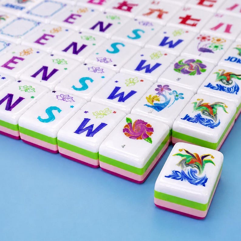 K&ouml;nnte beinhalten: Ein Set rechteckiger Mahjong-Spielsteine mit wei&szlig;en Oberseiten, die bunte Buchstaben E, N, S, W, florale Motive und chinesische Schriftzeichen zeigen. Jeder Spielstein hat eine geschichtete Basis mit gr&uuml;nen und rosa Streifen, angeordnet in einem Raster.