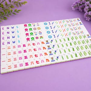 K&ouml;nnte beinhalten: Ein Set aus wei&szlig;en Mahjong-Spielsteinen, die in einem Raster angeordnet sind und bunte Druckmotive wie chinesische Schriftzeichen, Blumenmuster, Bambusstangen, Kreise, die Buchstaben E, N, W, S sowie mit Joker beschriftete Steine zeigen.