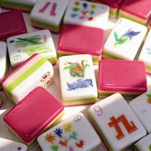 K&ouml;nnte beinhalten: Eine Sammlung rechteckiger Mahjong-Spielsteine mit einem geschichteten Design in Wei&szlig;, Hellgr&uuml;n und Dunkelrosa. Die Spielsteine zeigen verschiedene bunte, aufgemalte Motive, darunter Blumenmuster, numerische Zeichen und abstrakte Symbole.