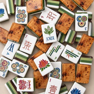 May include: A collection of Mahjong tiles featuring a mix of designs, including tiles with brown wood-grain backs, green striped patterns, and white faces displaying various symbols, numbers, floral motifs, a Joker label, and the text CRAK.