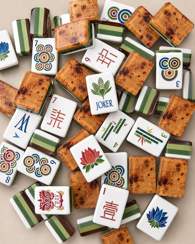 May include: A collection of Mahjong tiles featuring a mix of designs, including tiles with brown wood-grain backs, green striped patterns, and white faces displaying various symbols, numbers, floral motifs, a Joker label, and the text CRAK.