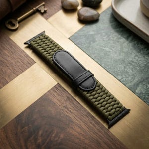 Könnte beinhalten: Ein olivgrünes, geflochtenes Uhrenarmband mit einer schwarzen, lederartigen Verstelllasche und schwarzen Metallanschlüssen an beiden Enden. Das Armband weist ein strukturiertes, gewebtes Muster auf und dient zur Befestigung einer Digitaluhr.