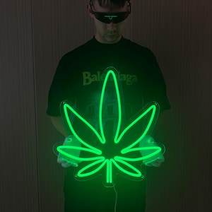 Puede incluir: Un letrero de neón verde brillante con forma de hoja de cannabis. El letrero presenta un contorno de luz verde brillante continua en forma de hoja de siete puntas. El tubo de neón está montado sobre una base de acrílico transparente.