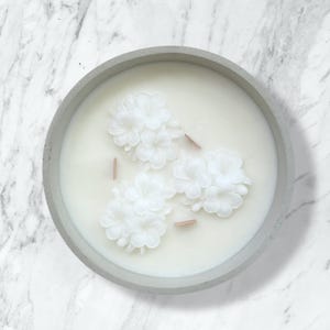 Puede incluir: Una vela redonda en un recipiente de hormigón gris relleno de cera blanca. La superficie presenta tres grupos de decoraciones de cera con forma de flor blanca y tres mechas de madera finas y planas dispuestas en triángulo en la cera.