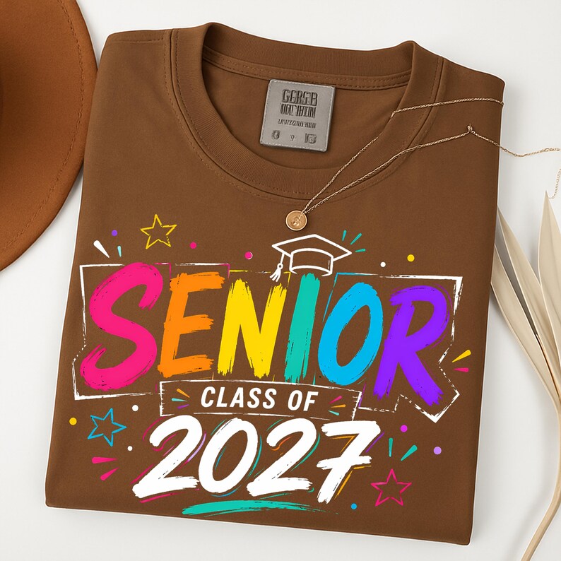 May include: A brown t-shirt featuring a colorful graphic with the text SENIOR CLASS OF 2027. The design includes a white graduation cap icon, multi-colored brushstroke lettering, and small star shapes in pink, yellow, and blue against the brown fabric.