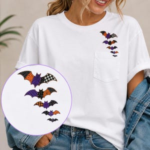 Op de afbeelding: Een wit T-shirt met korte mouwen en een borstzak. Boven de zak bevindt zich een verticaal grafisch ontwerp met vijf vleermuizen in afnemende grootte. De vleermuizen hebben patronen in oranje, paars, zwart en wit, waaronder stippen en ruitjes.