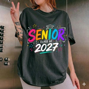 May include: A charcoal gray short-sleeve t-shirt featuring a colorful graphic print that reads SENIOR CLASS OF 2027 in bold, multi-colored lettering. The design includes a white graduation cap icon and small star accents in pink, yellow, and blue.