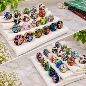 May include: A collection of round ceramic cabinet knobs featuring various hand-painted floral and geometric patterns in vibrant colors including red, blue, yellow, green, pink, and black, arranged on white folded towels.