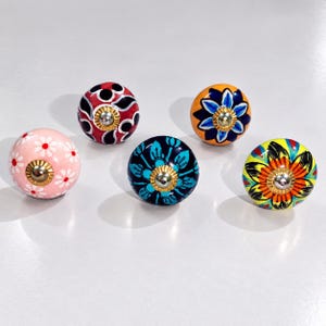 May include: Five round ceramic cabinet knobs featuring distinct hand-painted designs. One is light pink with white daisies, one is red with black and white patterns, one is orange with a blue flower, one is dark blue with teal leaves, and one is yellow.