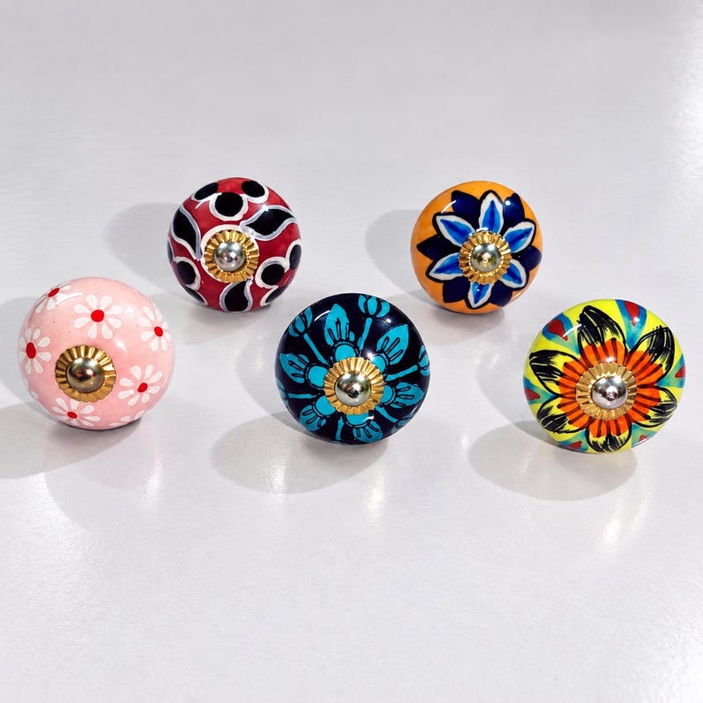 May include: Five round ceramic cabinet knobs featuring distinct hand-painted designs. One is light pink with white daisies, one is red with black and white patterns, one is orange with a blue flower, one is dark blue with teal leaves, and one is yellow.