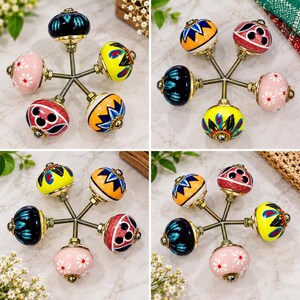 May include: A set of five round ceramic cabinet knobs featuring diverse patterns and colors, including pink with white flowers, yellow with blue and red accents, black with blue leaves, and red with black circular motifs, all with brass hardware.