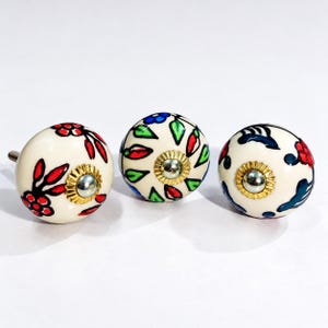 May include: Three round ceramic cabinet knobs with a cream-colored base. Each knob features hand-painted floral and leaf patterns in red, blue, and green. A metallic gold-toned circular plate with a central screw is visible on the front of each knob.