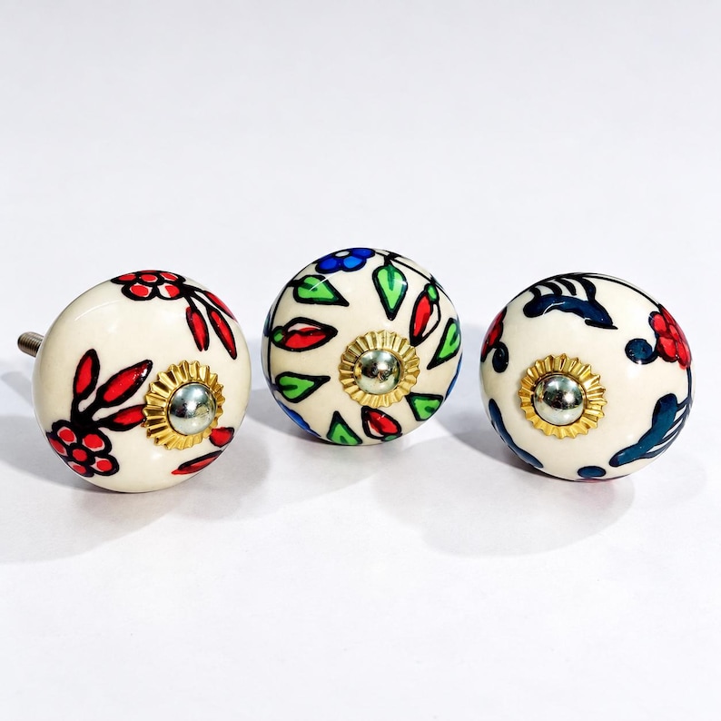 May include: Three round ceramic cabinet knobs with a cream-colored base. Each knob features hand-painted floral and leaf patterns in red, blue, and green. A metallic gold-toned circular plate with a central screw is visible on the front of each knob.