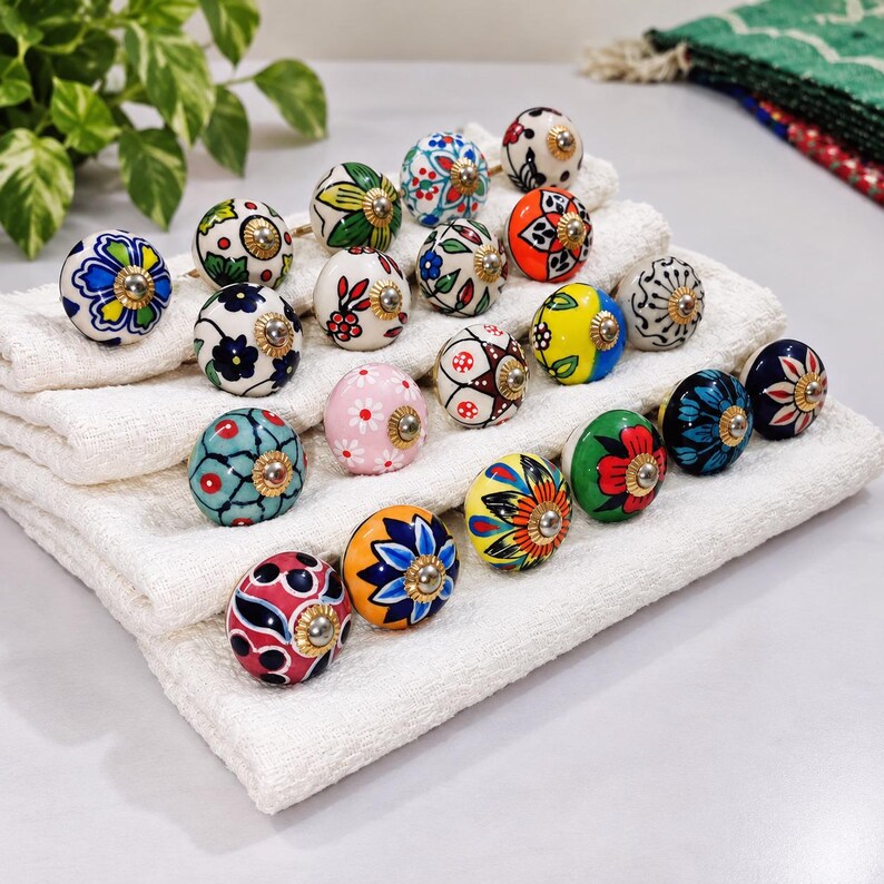 May include: A collection of round ceramic knobs featuring various hand-painted floral and geometric patterns in vibrant colors including red, blue, yellow, green, and pink. Each knob has a central metallic gold-tone hardware accent.