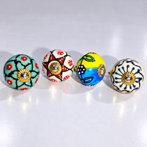 May include: Four round ceramic cabinet knobs with distinct hand-painted patterns. One is teal with red dots, one features a brown star with red dots, one is yellow and blue with floral motifs, and one is white with a dark blue radial line design.