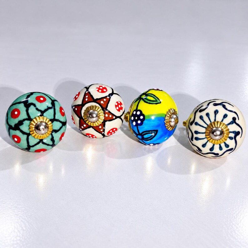 May include: Four round ceramic cabinet knobs with distinct hand-painted patterns. One is teal with red dots, one features a brown star with red dots, one is yellow and blue with floral motifs, and one is white with a dark blue radial line design.