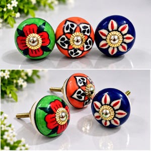 May include: Three round ceramic cabinet knobs featuring hand-painted floral patterns in green, orange, and blue. Each knob has a central gold-toned metal hardware piece with a fluted edge and a threaded metal screw extending from the back.