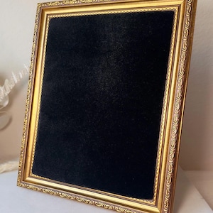 May include: A rectangular display board featuring a black velvet-textured surface framed by an ornate gold-colored border with intricate embossed patterns. The frame has a metallic finish and a raised edge, providing a contrast to the dark center.