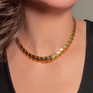 Puede incluir: Un collar dorado con un diseño geométrico texturizado. El collar se usa sobre una parte superior negra, creando un contraste. La pieza de joyería es de estilo gargantilla, cerca del cuello.