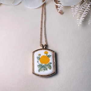 May include: A gold-coloured pendant necklace. The rectangular pendant has a white background with a yellow flower and green leaves. The delicate gold chain complements the pendant. The necklace is suitable for everyday wear.