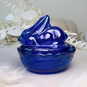 May include: A cobalt blue glass bunny figurine sitting in a matching basket-shaped dish. The bunny has long ears and is in a resting position. The glass is smooth and reflective, and the piece is likely a decorative item.