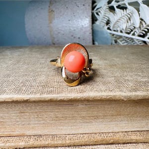 This vintage faux coral and gold Avon ring is a pretty little find. Gold tone ring featuring a vibrant faux coral stone in the center of an intricate gold arch loop. Major abstract space vibes! A unique statement piece by Avon from 1975. Signed in excellent vintage condition. 

Dimensions: US size 6-7 has inner sizer for flexible fit