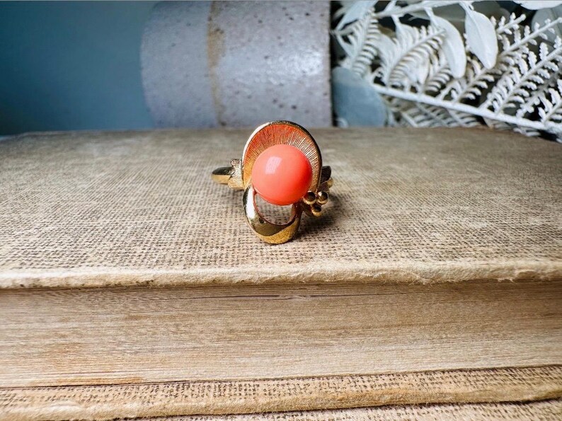 This vintage faux coral and gold Avon ring is a pretty little find. Gold tone ring featuring a vibrant faux coral stone in the center of an intricate gold arch loop. Major abstract space vibes! A unique statement piece by Avon from 1975. Signed in excellent vintage condition. 

Dimensions: US size 6-7 has inner sizer for flexible fit