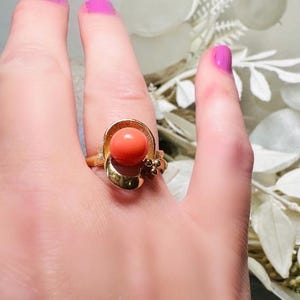 This vintage faux coral and gold Avon ring is a pretty little find. Gold tone ring featuring a vibrant faux coral stone in the center of an intricate gold arch loop. Major abstract space vibes! A unique statement piece by Avon from 1975. Signed in excellent vintage condition. 

Dimensions: US size 6-7 has inner sizer for flexible fit