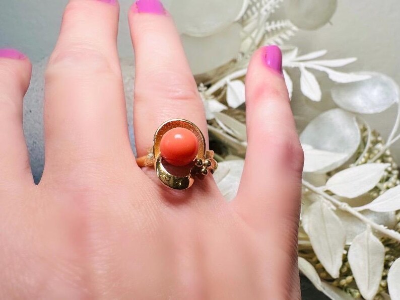 This vintage faux coral and gold Avon ring is a pretty little find. Gold tone ring featuring a vibrant faux coral stone in the center of an intricate gold arch loop. Major abstract space vibes! A unique statement piece by Avon from 1975. Signed in excellent vintage condition. 

Dimensions: US size 6-7 has inner sizer for flexible fit