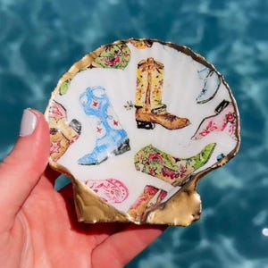 Op de afbeelding: Een witte sint-jakobsschelp met een herhalend patroon van kleurrijke cowboylaarzen in blauwe, gele, groene en roze tinten. De randen van de schelp zijn beschilderd met een metallic gouden afwerking, wat een decoratieve rand rond de print vormt.