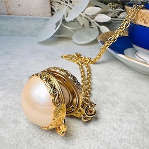Puede incluir: Un collar con colgante dorado con una gran esfera nacarada. El colgante tiene detalles ornamentados de estilo filigrana y una cadena a juego. El collar se exhibe cerca de una taza de té azul y blanca, lo que sugiere una estética vintage.