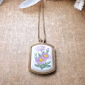May include: A gold-coloured rectangular pendant necklace with a painted floral design. The pendant has a white background with purple and yellow flowers and green leaves. The chain is a delicate gold colour.