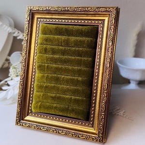 May include: A rectangular jewelry display stand featuring an ornate, antique-style gold frame with dark brown detailing. The interior is filled with a plush, olive green velvet insert containing eight horizontal slots designed for holding rings.