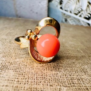 This vintage faux coral and gold Avon ring is a pretty little find. Gold tone ring featuring a vibrant faux coral stone in the center of an intricate gold arch loop. Major abstract space vibes! A unique statement piece by Avon from 1975. Signed in excellent vintage condition. 

Dimensions: US size 6-7 has inner sizer for flexible fit