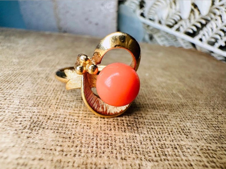 This vintage faux coral and gold Avon ring is a pretty little find. Gold tone ring featuring a vibrant faux coral stone in the center of an intricate gold arch loop. Major abstract space vibes! A unique statement piece by Avon from 1975. Signed in excellent vintage condition. 

Dimensions: US size 6-7 has inner sizer for flexible fit