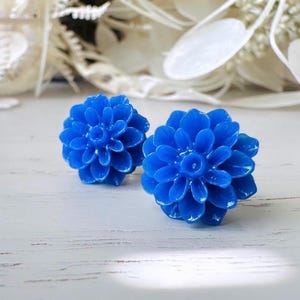 May include: Two vibrant blue flower-shaped stud earrings. The earrings have a detailed petal design and a glossy finish. They are set against a blurred background of white and beige decorative elements.