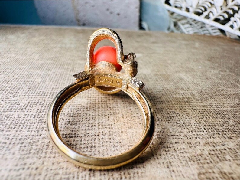 This vintage faux coral and gold Avon ring is a pretty little find. Gold tone ring featuring a vibrant faux coral stone in the center of an intricate gold arch loop. Major abstract space vibes! A unique statement piece by Avon from 1975. Signed in excellent vintage condition. 

Dimensions: US size 6-7 has inner sizer for flexible fit