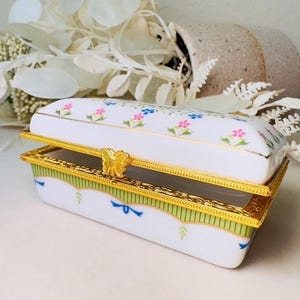 以下が含まれることがあります： 長方形の白い磁器製の小物入れ。蝶番式の蓋が付いています。蓋とベースは花柄と金色のトリムで装飾されています。金色の蝶の留め金が蓋を固定します。ボックスには緑と青のアクセントがあります。