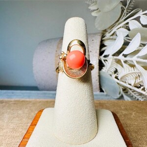 This vintage faux coral and gold Avon ring is a pretty little find. Gold tone ring featuring a vibrant faux coral stone in the center of an intricate gold arch loop. Major abstract space vibes! A unique statement piece by Avon from 1975. Signed in excellent vintage condition. 

Dimensions: US size 6-7 has inner sizer for flexible fit