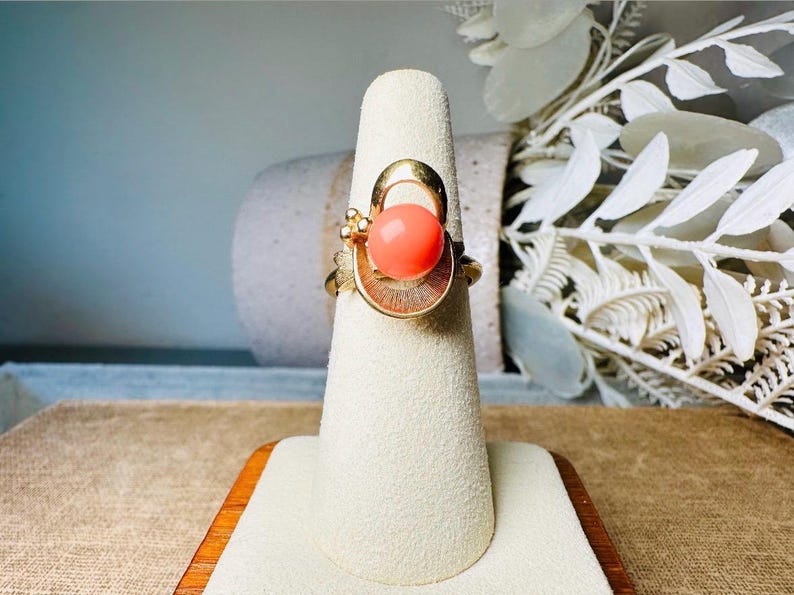 This vintage faux coral and gold Avon ring is a pretty little find. Gold tone ring featuring a vibrant faux coral stone in the center of an intricate gold arch loop. Major abstract space vibes! A unique statement piece by Avon from 1975. Signed in excellent vintage condition. 

Dimensions: US size 6-7 has inner sizer for flexible fit