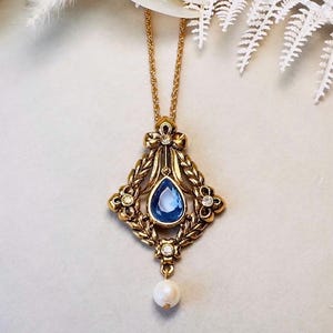 May include: Gold-coloured pendant necklace with a blue, teardrop-shaped stone at its centre. The pendant has an ornate design with floral and leaf accents, and a small pearl hangs below. The necklace chain is also gold-coloured.