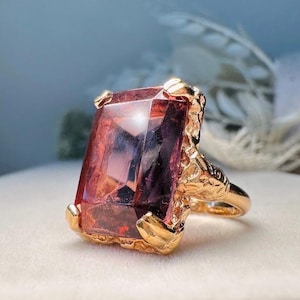 May include: A gold ring with a large, rectangular, reddish-purple gemstone. The ring has a textured gold band and setting, with claw-like prongs securing the stone. The gemstone reflects light, showcasing its facets.