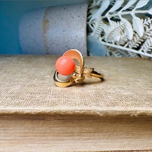 This vintage faux coral and gold Avon ring is a pretty little find. Gold tone ring featuring a vibrant faux coral stone in the center of an intricate gold arch loop. Major abstract space vibes! A unique statement piece by Avon from 1975. Signed in excellent vintage condition. 

Dimensions: US size 6-7 has inner sizer for flexible fit