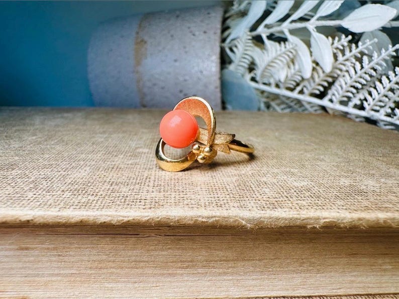 This vintage faux coral and gold Avon ring is a pretty little find. Gold tone ring featuring a vibrant faux coral stone in the center of an intricate gold arch loop. Major abstract space vibes! A unique statement piece by Avon from 1975. Signed in excellent vintage condition. 

Dimensions: US size 6-7 has inner sizer for flexible fit