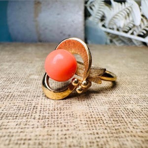 This vintage faux coral and gold Avon ring is a pretty little find. Gold tone ring featuring a vibrant faux coral stone in the center of an intricate gold arch loop. Major abstract space vibes! A unique statement piece by Avon from 1975. Signed in excellent vintage condition. 

Dimensions: US size 6-7 has inner sizer for flexible fit