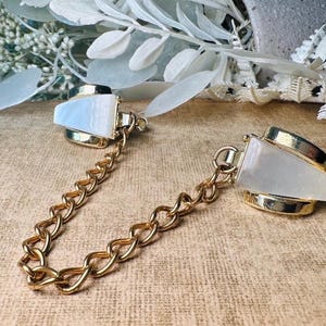 Op de afbeelding: Goudkleurige metalen kraagclips met een ketting. Elke clip heeft een rechthoekige, iriserende, witte steen in een gouden frame. De clips zijn verbonden door een gouden ketting, waardoor een stijlvol accessoire ontstaat.